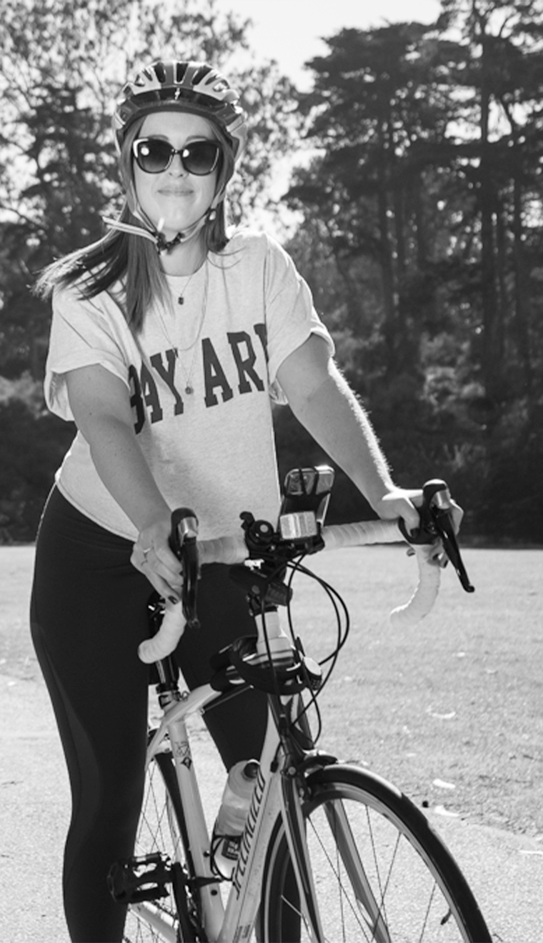 Liz, proud Bay Area Native. Putting her first since 2013. Woman in mid-20s straddles a bicycle. She wears sunglasses and a helmet. Her shirt reads