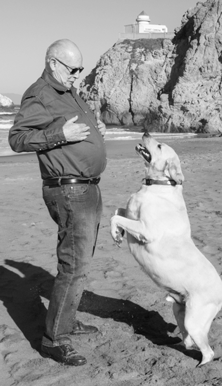 Dwayne, enjoying retirement with Gridley. Putting him first since 2019. Older man wearing sunglasses, and a button-up shirt embraces a dog on the beach.
